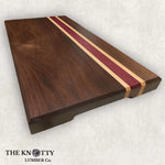 A rectangular cutting board made of walnut, maple, and purpleheart wood with a striped pattern, branded with 'The Knotty Lumber Co.'.