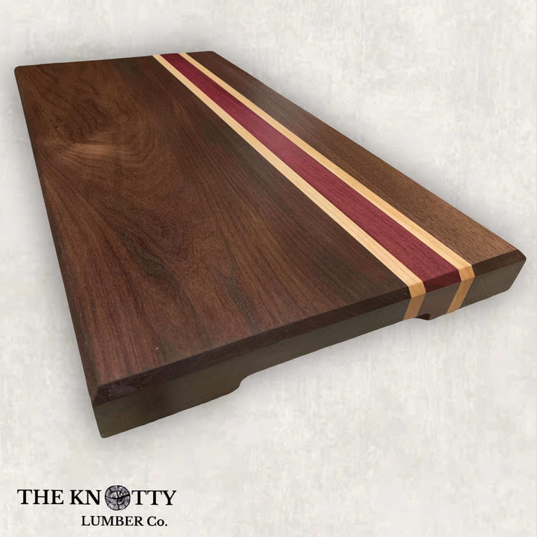 A rectangular cutting board made of walnut, maple, and purpleheart wood with a striped pattern, branded with 'The Knotty Lumber Co.'.
