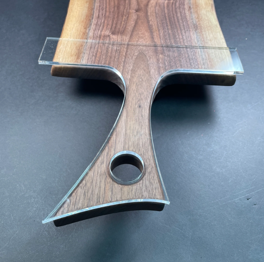 A close-up of a wooden serving board or cutting board with a clear acrylic guide or insert placed along the top edge, photographed on a dark gray surface.