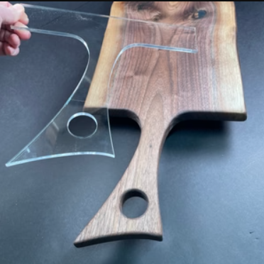 A clear acrylic cutting board guide or template with a circular hole, shown being held over a wooden serving board on a dark surface.
