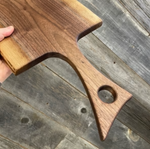 A close-up of a hand holding a wooden serving board or cutting board with a handle cutout, shown against rustic weathered wood planks.