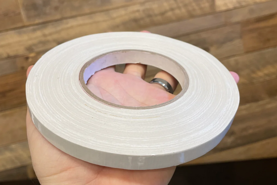 A hand holding a roll of white adhesive tape with a cardboard core, set against a rustic wooden plank wall background.