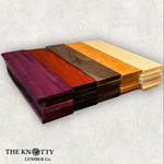 A variety of rough cut lumber pieces in different colors, including Purpleheart, Padauk, Hard Maple, Black Walnut, and Black Cherry, arranged side by side.